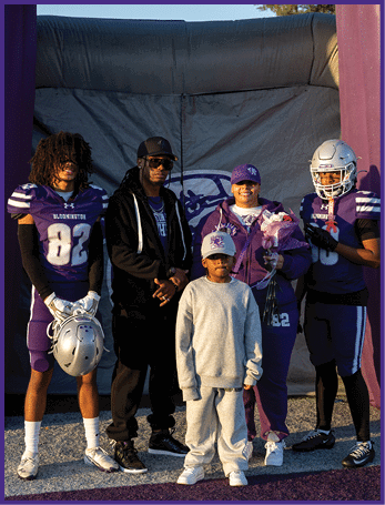 Five people stand together on a sports field entrance, with two wearing football uniforms holding helmets, one holding flowers, and two others dressed casually. Description generated by AI