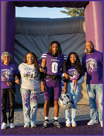Two people in purple football uniforms stand with three casually dressed adults smiling outdoors in front of an inflatable backdrop. Description generated by AI
