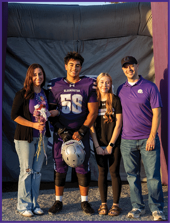 Four people stand together smiling in front of a large inflatable structure, with one person wearing a football uniform holding a helmet and the others dressed casually. Description generated by AI