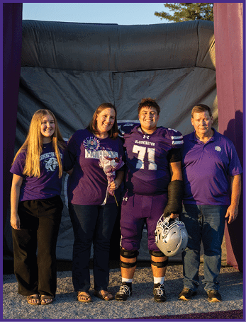 Four people stand together smiling under a large inflatable arch, with one person wearing a football uniform holding a helmet and another holding a bouquet of flowers. Description generated by AI