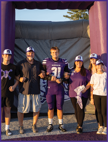 Five people stand arm in arm in front of an inflatable sports tunnel, with one person wearing a purple football uniform and the others wearing casual clothing and matching hats. Description generated by AI