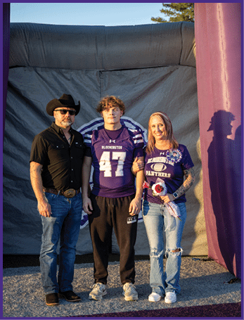 Three people stand together outdoors in front of a large inflatable structure, with one person wearing a sports jersey and the others dressed casually, one holding a bouquet of flowers. Description generated by AI