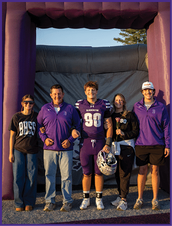 Five people stand arm in arm under a large purple inflatable arch, with one person wearing a football uniform holding a helmet and others dressed casually in sports-themed clothing, all smiling in an outdoor setting. Description generated by AI