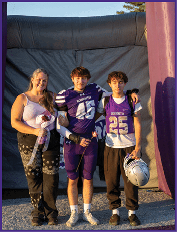 Three people stand together smiling under a large purple inflatable arch, with two wearing purple football uniforms and one holding flowers. Description generated by AI