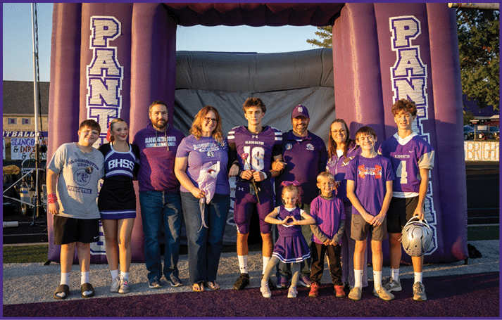 A group of people including children and adults wearing purple sports and casual clothing stand together smiling in front of a large inflatable structure with the word “PANTHERS" on it at an outdoor event. Description generated by AI