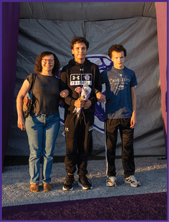 Three people stand arm in arm on a track with a purple backdrop, one holding a bouquet of flowers and all dressed casually in athletic and casual clothing. Description generated by AI