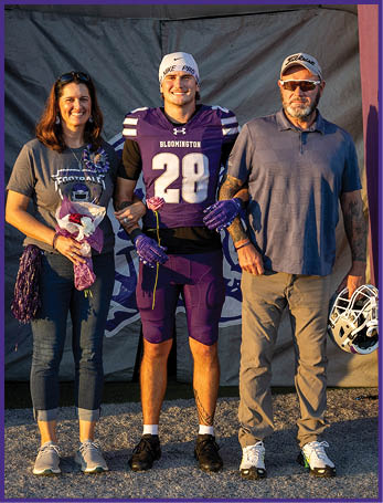 Three people stand together smiling with one person in a purple football uniform holding a flower and the other two casually dressed, one holding a football helmet. Description generated by AI