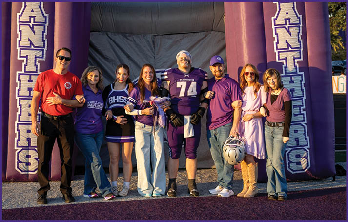 A group of people dressed in casual and sports attire stand together smiling under a large purple inflatable arch on a sports field during sunset. Description generated by AI
