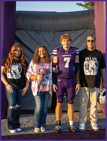 Four people stand side by side outdoors with one person wearing a purple football uniform holding a helmet and the others wearing casual clothing including matching black shirts with a graphic design. Description generated by AI
