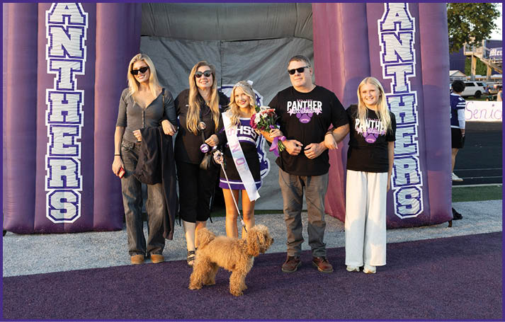 Five people standing together on a purple carpet with one person wearing a sash and holding flowers, accompanied by a small brown dog, in front of a large purple inflatable arch. Description generated by AI
