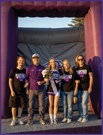 Five people stand together smiling outdoors with one person in a cheerleading outfit holding flowers and a sash while the others wear matching Panther-themed shirts. Description generated by AI