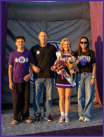 Four people stand together outdoors with one person in a cheerleading outfit holding flowers while the others wear casual clothing and stand under a large inflatable structure. Description generated by AI