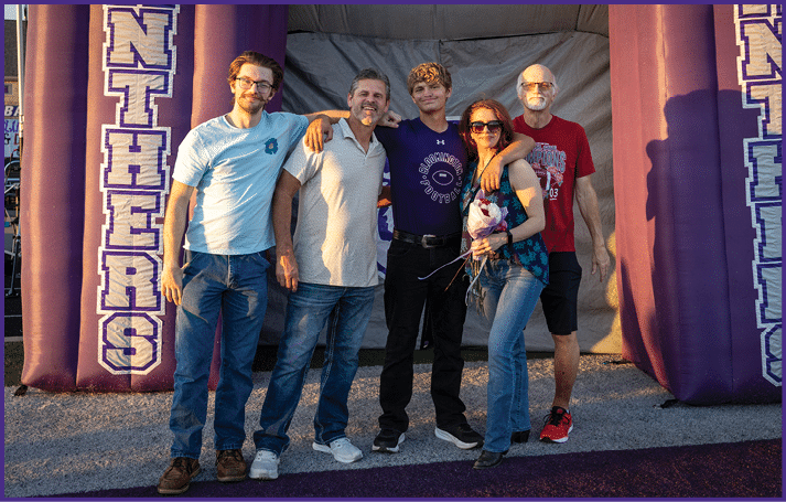 Five adults stand closely together outdoors with arms around each other, smiling and posing for a photo, with one holding a bouquet of flowers. Description generated by AI