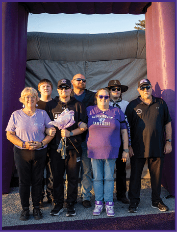 A group of seven people standing together outdoors on a paved surface with a dark backdrop, some wearing sunglasses and casual clothing including a purple shirt with “Bloomington Panthers" text, and one person holding a bouquet of flowers. Description generated by AI