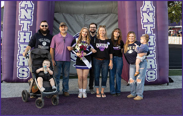 A group of seven people, including one holding a child and one pushing a stroller with a child, stand together smiling in front of a large inflatable arch with the word “Panthers" on it, with one person dressed in a cheerleader outfit holding flowers and wearing a sash. Description generated by AI