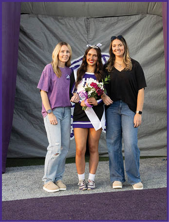 Three adults stand together smiling on a purple carpeted area with one wearing a cheerleading outfit and holding flowers and a sash. Description generated by AI
