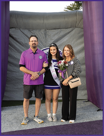 Three people stand together on a sports field with one person in a cheerleading outfit and sash holding flowers, flanked by two others wearing casual clothing. Description generated by AI