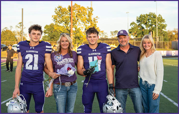 Two people in football uniforms holding helmets stand on a field with three casually dressed people between them, all smiling and posing for a photo during daylight. Description generated by AI