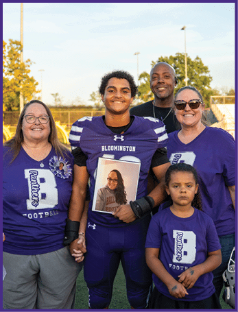 A person in a football uniform stands on a field holding hands with two people wearing matching purple shirts, while two other people stand behind them smiling, with one holding a bouquet of flowers. Description generated by AI