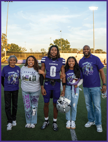 Five people stand arm in arm on a football field at sunset with one person wearing a football uniform and holding a helmet while others wear senior-themed shirts and casual clothing. Description generated by AI