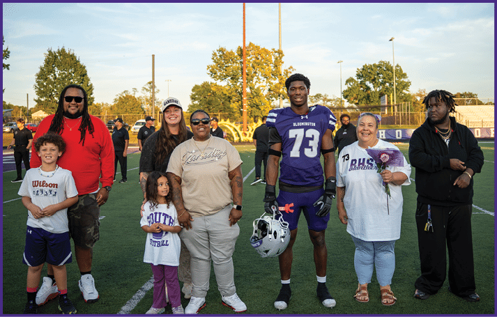 A group of people stand together on a sports field with one person in a purple football uniform holding a helmet and others wearing casual clothing, some smiling and one holding a flower. Description generated by AI