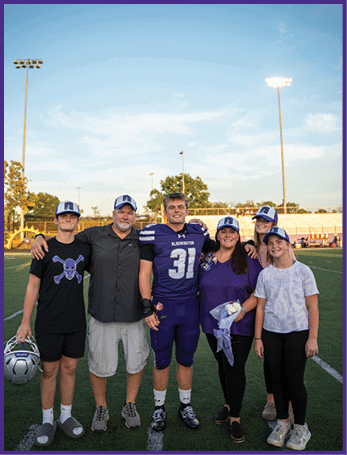 A group of six people stands together on a sports field with one person in a football uniform holding a flower, all wearing matching hats and smiling. Description generated by AI