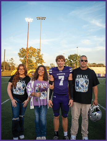 Four people stand on a sports field at sunset with one person wearing a purple football uniform and the others wearing matching black and purple shirts, with one holding a flower. Description generated by AI