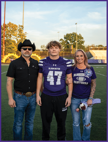 Three people stand arm in arm on a football field at sunset, with one person wearing a purple Bloomington football jersey and the others dressed casually, one in a black shirt and cowboy hat and the other in a purple Bloomington Panthers shirt holding flowers. Description generated by AI