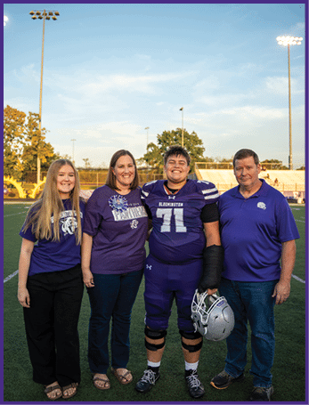 Four people stand together on a sports field with one person wearing a football uniform and holding a helmet, all smiling and dressed in casual or team-related clothing. Description generated by AI