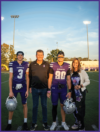 Two people in purple football uniforms holding helmets stand next to two adults on a football field under a clear sky with stadium lights. Description generated by AI