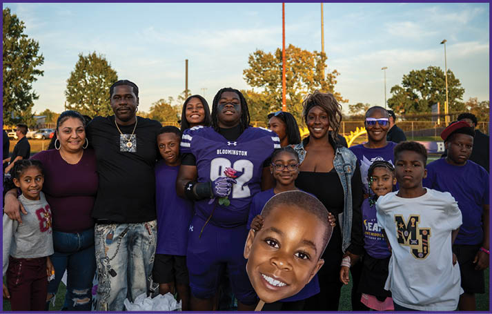 A group of people, including one in a football uniform holding a flower, stand closely together outdoors with trees and a sports field in the background, and one person holds a large cutout of a smiling child's face. Description generated by AI