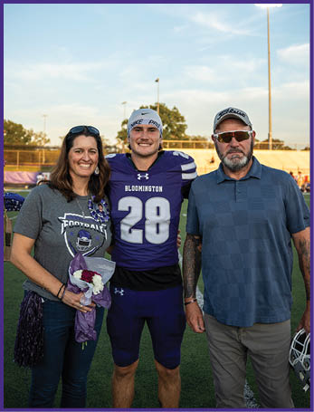Three people stand on a football field at sunset with one person in a purple football uniform holding flowers and the other two wearing casual clothing and sunglasses. Description generated by AI