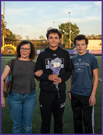 Three people stand on a sports field at sunset with one person in the center holding a bouquet of flowers and the others standing closely on either side. Description generated by AI