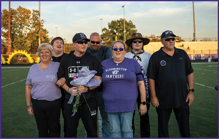 A group of seven people standing together on a sports field at sunset with some wearing casual clothing and sunglasses, and one person holding a bouquet of flowers. Description generated by AI
