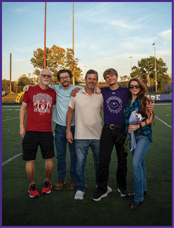 Five people stand closely together on a sports field at sunset with trees and stadium lights in the background, smiling and posing for the photo. Description generated by AI