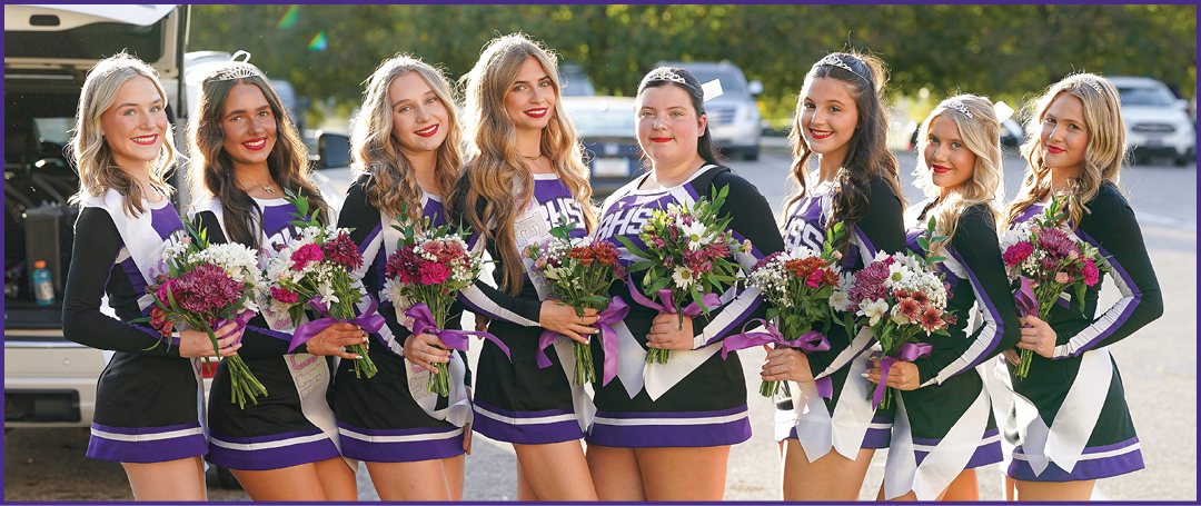 Six people in matching cheerleading uniforms holding bouquets of flowers stand closely together outdoors with cars and trees in the background. Description generated by AI