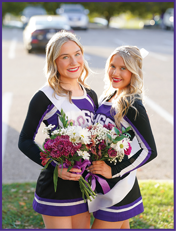 Two adults wearing purple and white cheerleading uniforms smile outdoors in bright sunlight. Description generated by AI