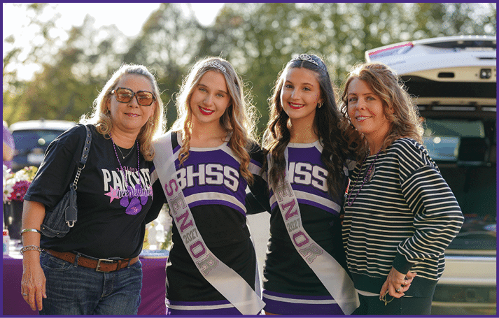 Two people wearing cheerleading uniforms with “RHSS" and "Senior 2021" sashes stand smiling outdoors next to a person in casual clothing and sunglasses. Description generated by AI