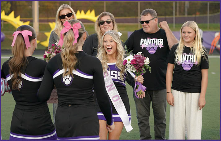 A group of people on a sports field includes a person wearing a sash and holding flowers smiling at two others in cheerleading uniforms with pink bows, while three others stand nearby wearing matching black shirts with “Panther" text. Description generated by AI