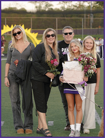 Five people standing on a sports field with one person holding a basket of flowers and a bouquet, all wearing casual clothing and sunglasses. Description generated by AI