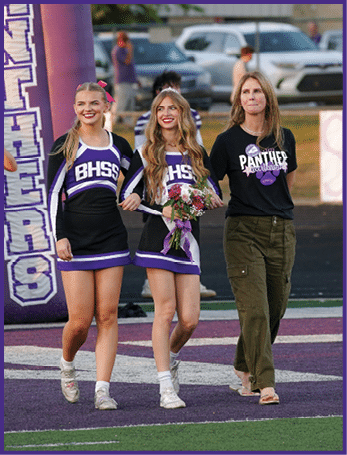 Two people in cheerleading uniforms walk arm in arm on a sports field alongside a person wearing casual clothing with a Panthers logo on the shirt. Description generated by AI