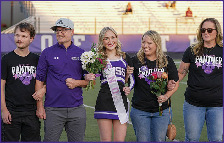 A group of people standing outdoors on a field with one person wearing a cheerleading outfit and sash holding flowers while others stand beside them smiling and holding flowers. Description generated by AI