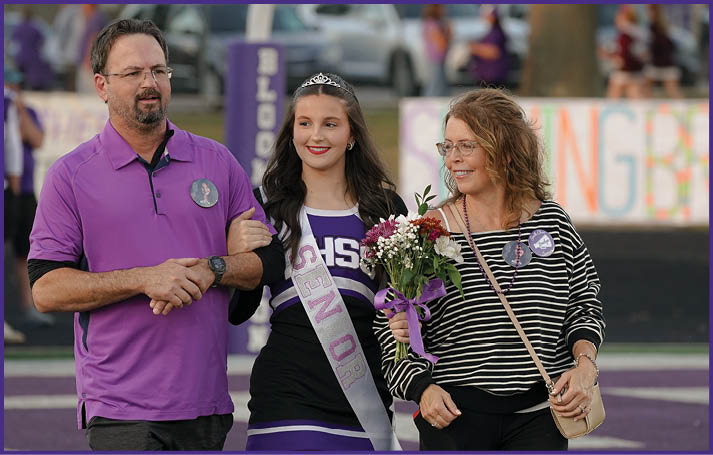 A person wearing a purple shirt walks arm-in-arm with a person in a cheerleading outfit and sash, while another person in a striped shirt and glasses walks beside them holding flowers on a sports field. Description generated by AI