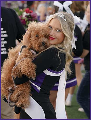 A person with long blonde hair wearing a tiara and a black outfit is smiling and hugging a curly-haired brown dog. Description generated by AI