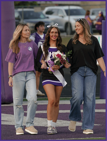 Three people walk together on a sports field with one person in the center wearing a cheerleading uniform, a sash, and holding flowers while the others wear casual clothing. Description generated by AI