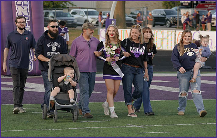 A group of people including a person in a cheerleader outfit holding flowers, others wearing casual and sports-themed clothing, and a child in a stroller are walking together on a sports field. Description generated by AI