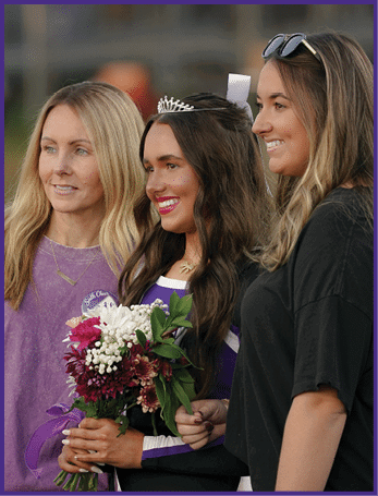 A person wearing a tiara and holding a bouquet of flowers stands smiling between two other people, one wearing a purple shirt and the other in black. Description generated by AI