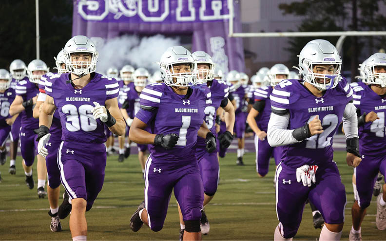 A group of people wearing purple football uniforms and helmets are running onto a field with a large purple banner and smoke in the background. Description generated by AI