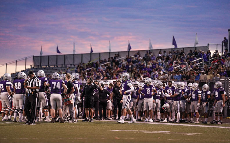 A group of people in football uniforms and coaches are gathered on a field near the sidelines with a crowd of spectators in the background under a colorful evening sky. Description generated by AI