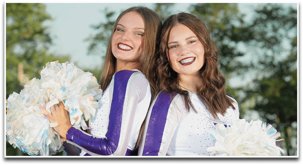 Two smiling adults with long hair and braces pose closely together outdoors with trees in the background. Description generated by AI